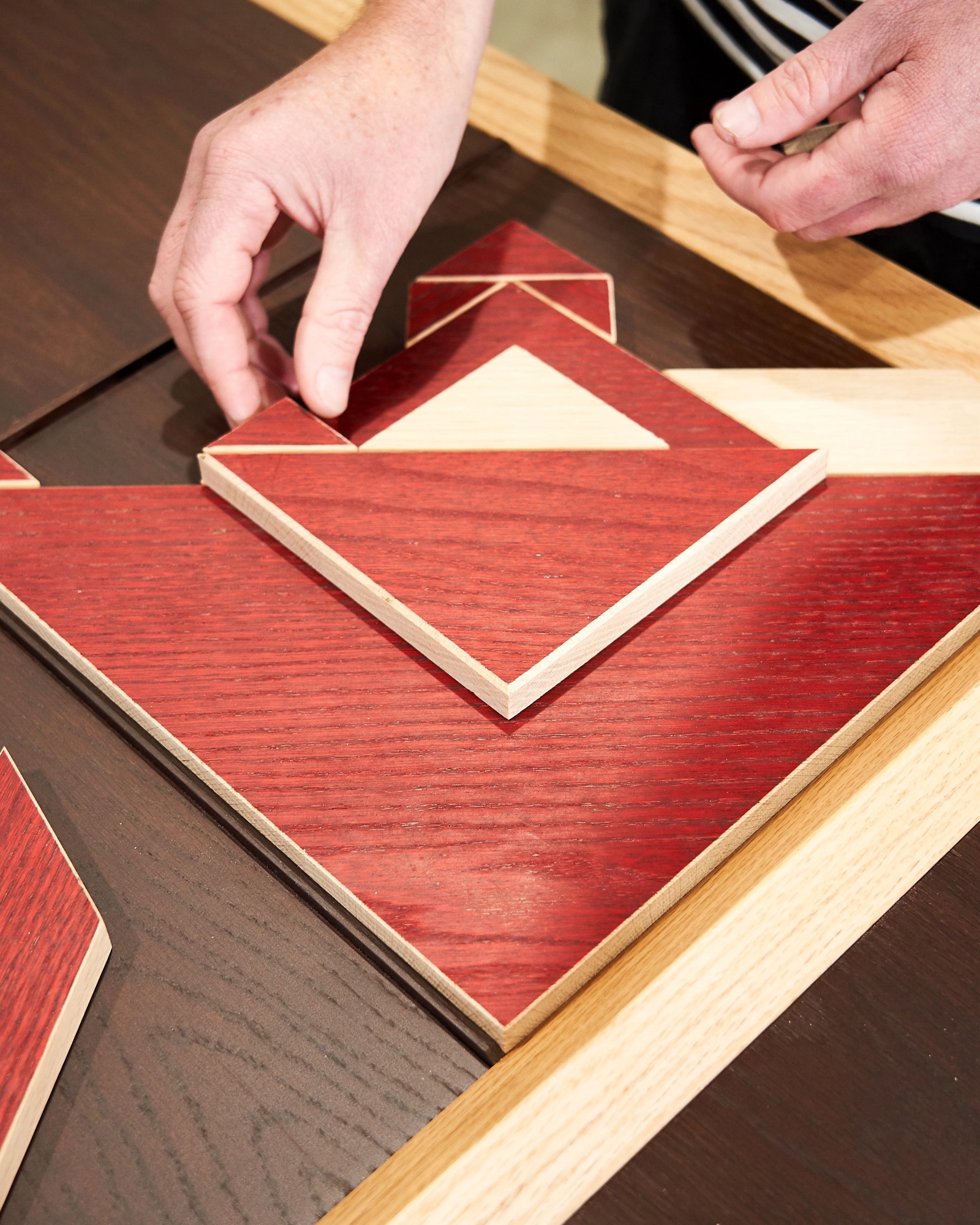 A hand placing red-stained American hardwood tangram tiles into a timber frame